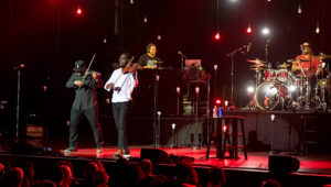 Black Violin two string musicians on stage playing string instruments with a dj and lights in the background.