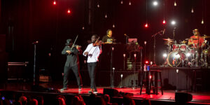 Black Violin two string musicians on stage playing string instruments with a dj and lights in the background.