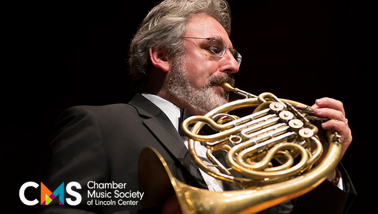 Radovan Vlatkovics, a man with gray hair and a beard, plays the French horn.