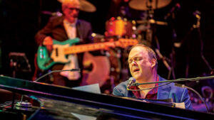 Rob Mathes sings at the piano, bass player in background.