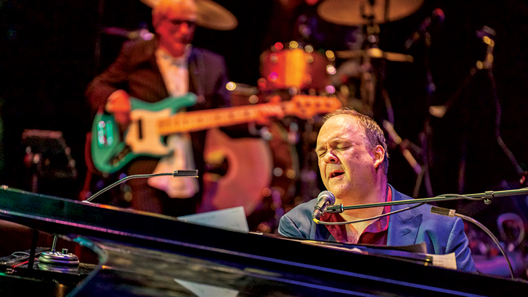 Rob Mathes sings at the piano, bass player in background.
