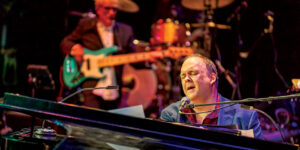 Rob Mathes sings at the piano, bass player in background.