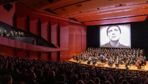 Voices of Light performance. Black and white film plays on a screen behind an orchestra.